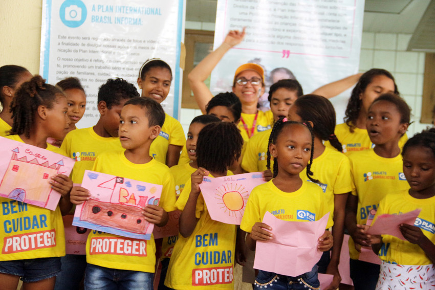 Crianças do projeto Cambalhotas, da Plan International Brasil, posam para a foto segurando os desenhos que fizeram. Entre elas, há também uma educadora da primeira infância. O clima é de felicidade.