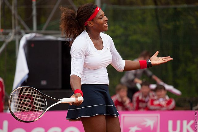 Tenista Serena Williams na quadra de tênis. Ela segura uma raquete na mão direita e acena de braços abertos na quadra.