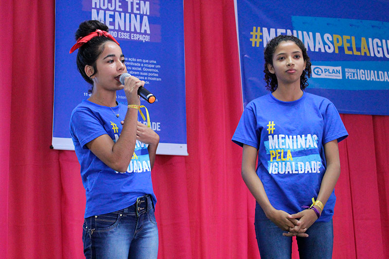 Duas meninas adolescentes estão em um palco, com fundo vermelho. Elas vestem uma camiseta azul com a frase "Meninas pela Igualdade", da campanha da Plan International Brasil. A menina da esquerda segura um microfone.