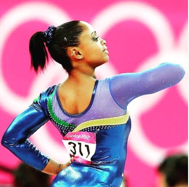 A ginasta Daiane dos Santos, mulher jovem negra, está apresentando seus movimentos, em pose imponente. Ela está de costas e veste um colant azul.