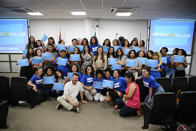 Meninas adolescentes posam para foto com educadores e apoiadores do projeto Escola de Liderança para Meninas, da Plan International Brasil. Elas seguram o certificado de formatura que receberam no evento.
