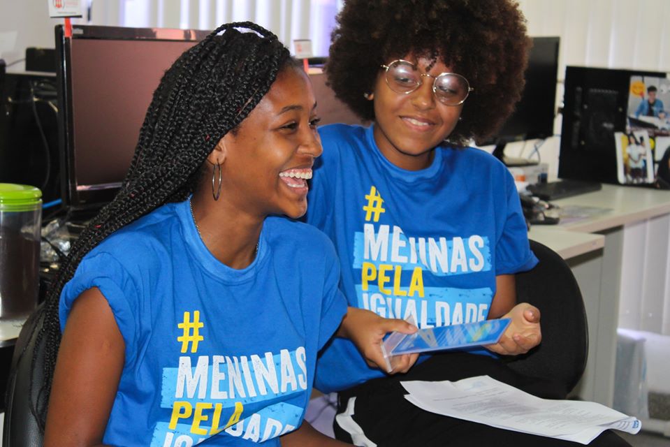 Duas meninas jovens negras encontram-se em um escritório. Elas estão sentadas e o clima é de descontração. Elas vestem uma camiseta azul com os dizeres Meninas pela Igualdade.