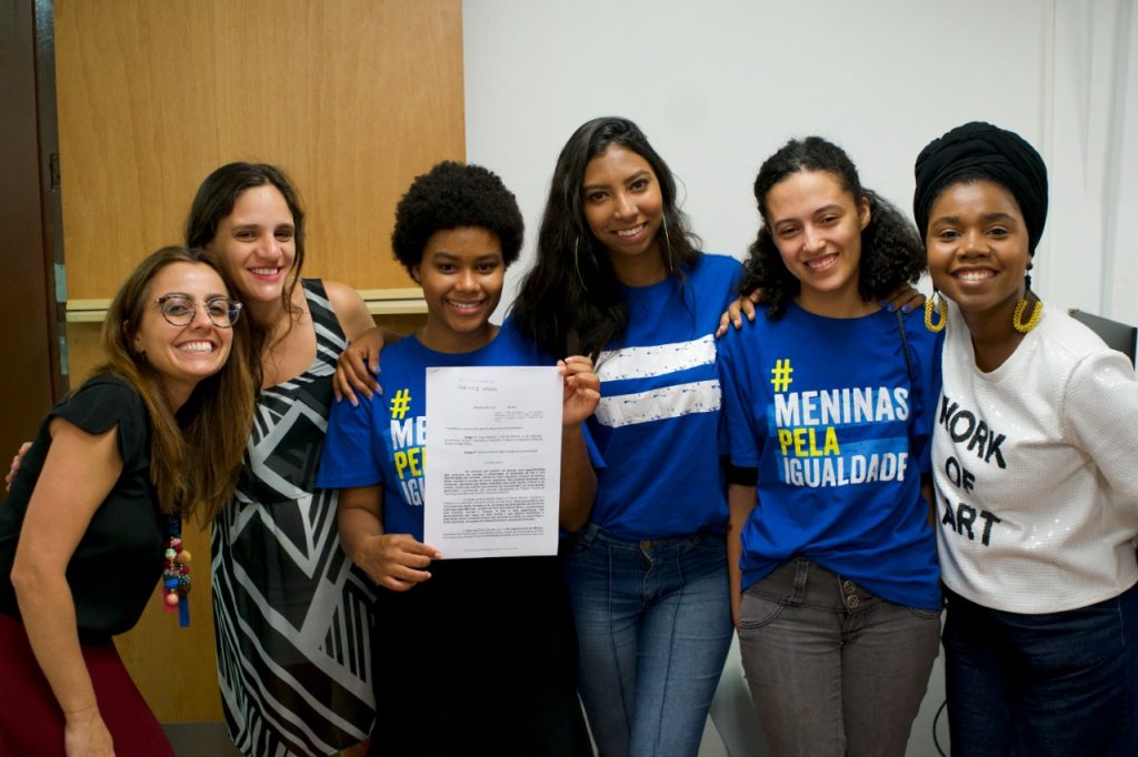 Meninas adolescentes posam entre três mulheres, em uma sala da Assembleia Legislativa de São Paulo. Uma das adolescentes segura o documento do projeto de lei que institui o dia da menina em São Paulo. Todas estão alegres e sorridentes.