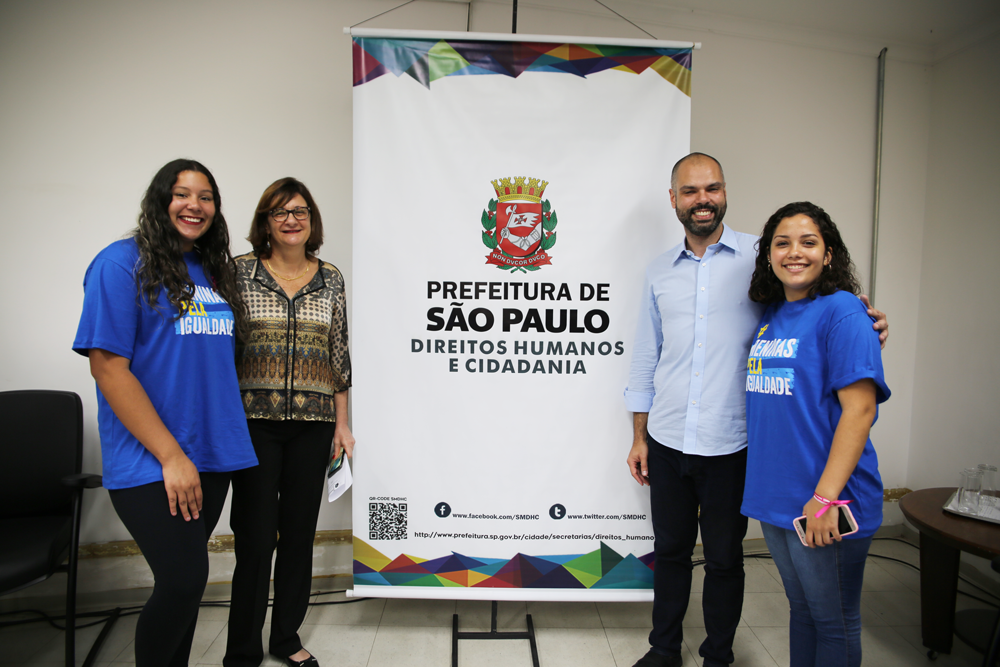 Meninas adolescentes posam com o prefeito de São Paulo e a secretária de Direitos Humanos de São Paulo. O clima é de alegria.