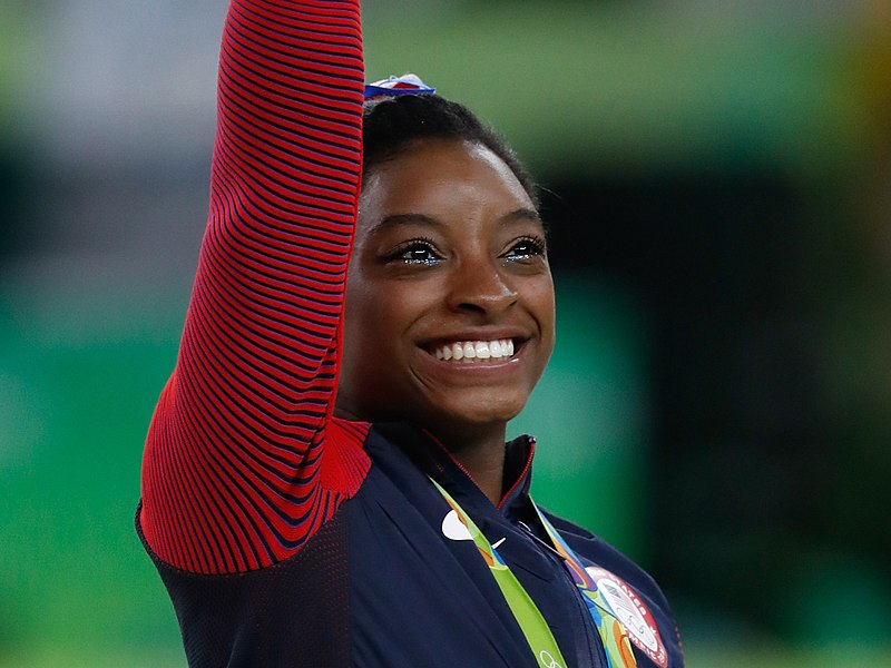 Simone Biles, atleta de ginástica artística e mulher jovem negra, está acenando em clima alegre.
