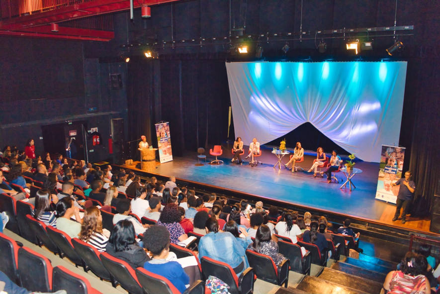 Auditório do teatro onde ocorreu o III Seminário da Primeira Infância. O público está sentado em cadeiras vermelhas olhando para o palco, onde os palestrantes da mesa de abertura dialogam sentados em cadeiras vermelhas.