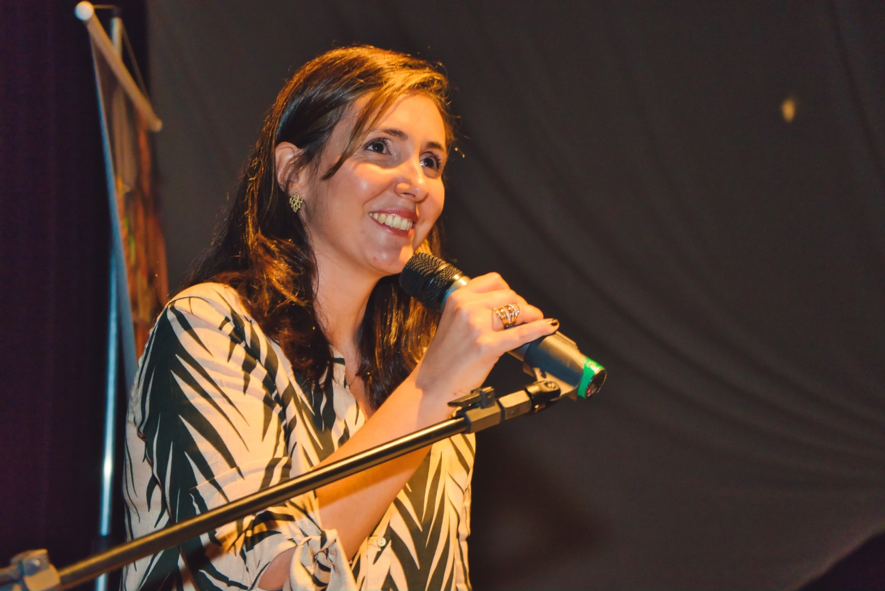 Uma mulher branca, de cabelos lisos, com uma roupa com estampas diagonais nas cores branco e azul escuro sorri ao segurar um microfone.