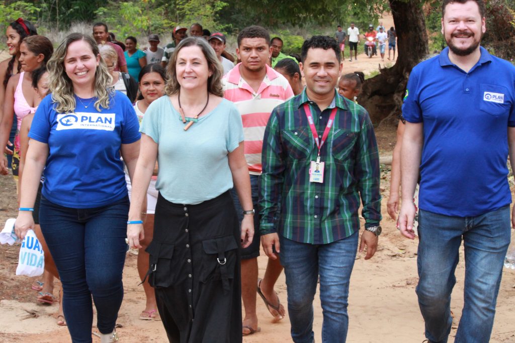 Duas mulheres e dois homens caminham lado a lado. Elas e eles fazem parte da equipe da Plan International Brasil, que luta pelos direitos das meninas. Atrás deles, há algumas pessoas caminhando também. Todos sorriem.