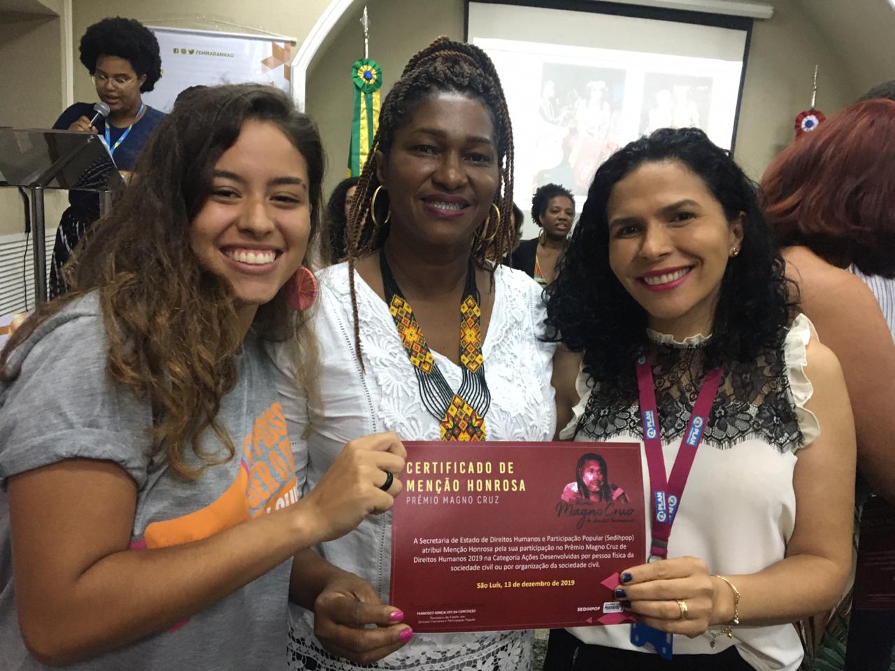 Uma jovem e duas mulheres posam segurando um certificado de menção honrosa do Prêmio Magno Luz concedido à Plan International Brasil. O clima é de alegria.