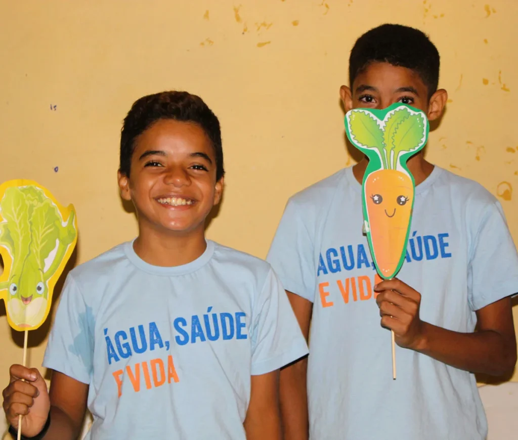 Dois meninos sorrindo seguram figuras de vegetais enquanto participam de uma atividade educativa sobre saúde e alimentação.