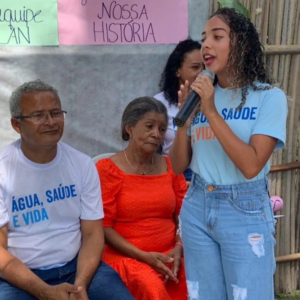 Jovem mulher discursa ao lado de pessoas da comunidade, promovendo debates sobre saúde, identidade e empoderamento feminino.