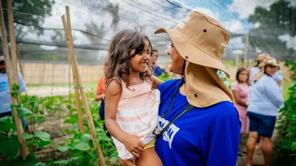 Mulher com chapéu da Plan International segurando e sorrindo para uma menina em uma plantação, representando ações de apadrinhamento infantil.