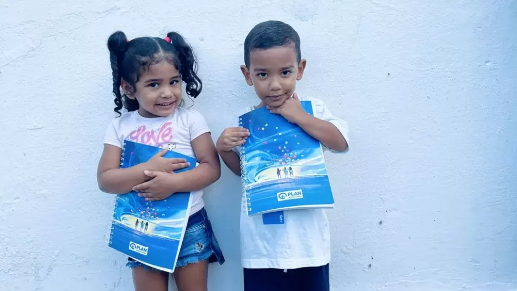 Menina e menino segurando cadernos da Plan International Brasil, sorrindo em frente a uma parede clara, representando os impactos positivos da doação mensal.