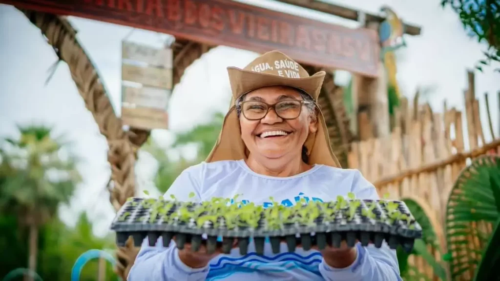 Mulher sorridente segurando bandeja de mudas em projeto da Plan International Brasil, simbolizando ações contra a pobreza e a violência.
