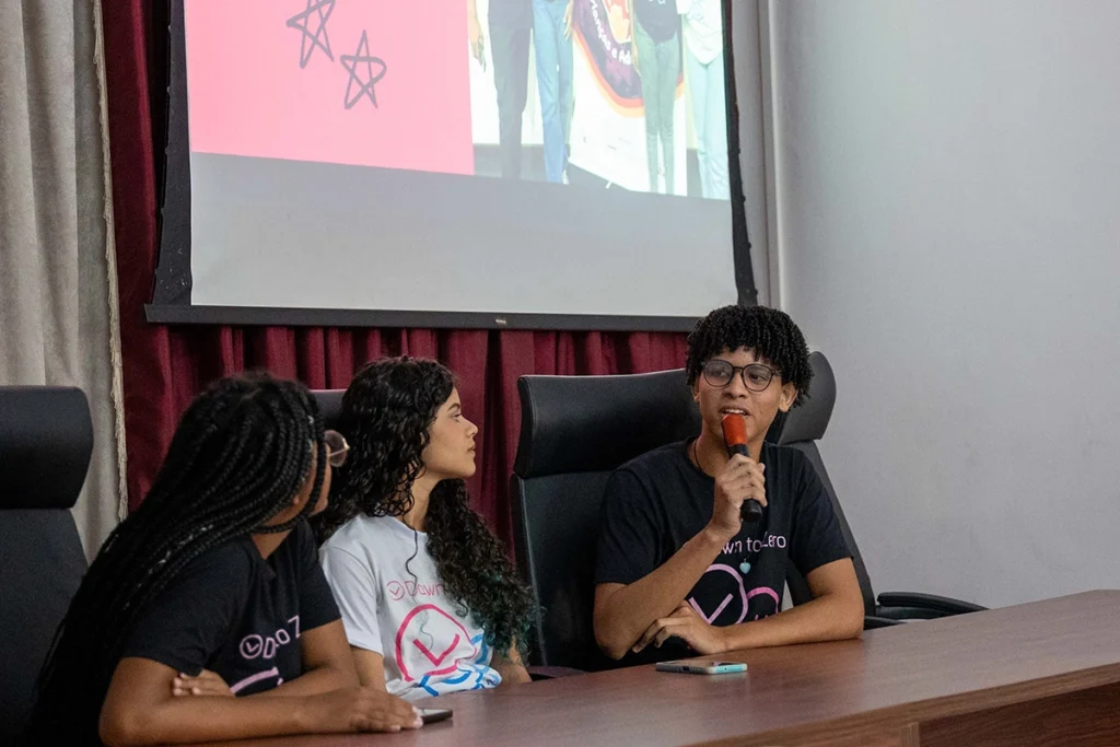 Jovens falam com o público em auditório, usando camisetas do projeto Down To Zero, em ação de conscientização sobre direitos e segurança de crianças e adolescentes.