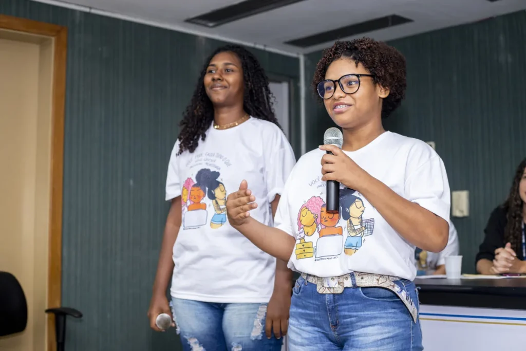 Duas jovens fazem apresentação com microfone, usando camisetas ilustradas, durante atividade de liderança e empoderamento feminino.