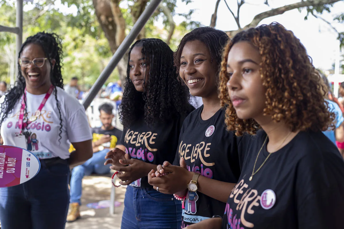 Quatro jovens mulheres participam de atividade em grupo ao ar livre, vestindo camisetas da campanha, sorrindo e interagindo de forma acolhedora.
