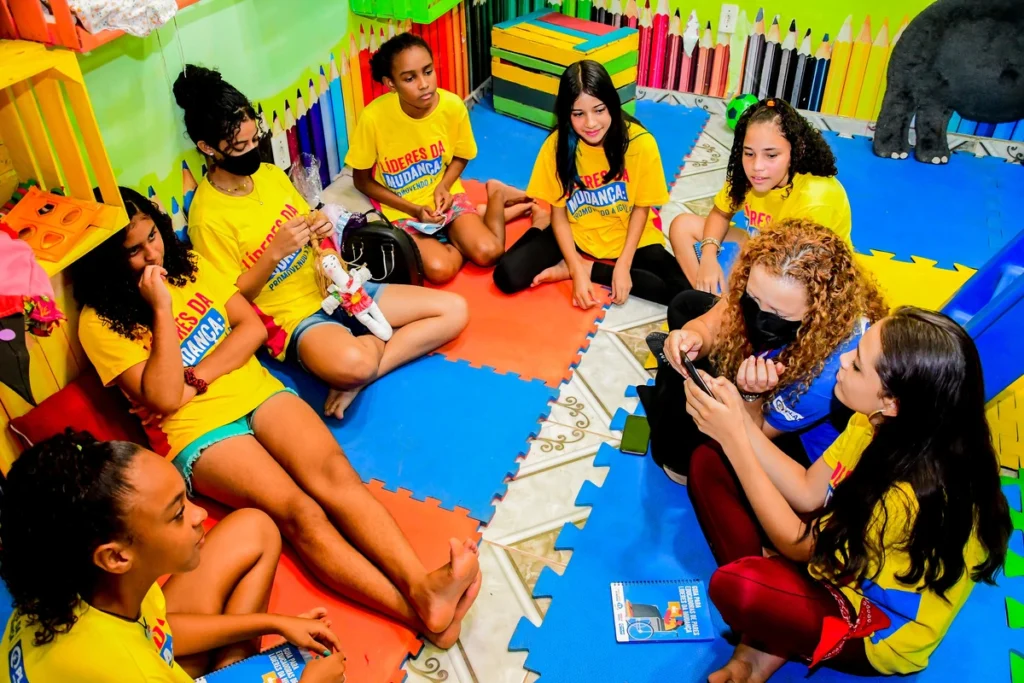 Grupo de meninas sentadas em roda, vestindo camisetas amarelas do Projeto Líderes da Mudança, interagindo em ambiente educativo e colorido.