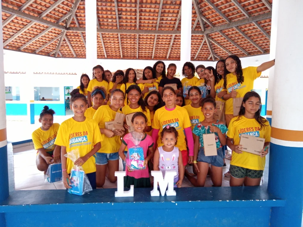 Grupo de meninas de diferentes idades posa para foto de celebração, segurando materiais educativos, em evento do Projeto Líderes da Mudança.