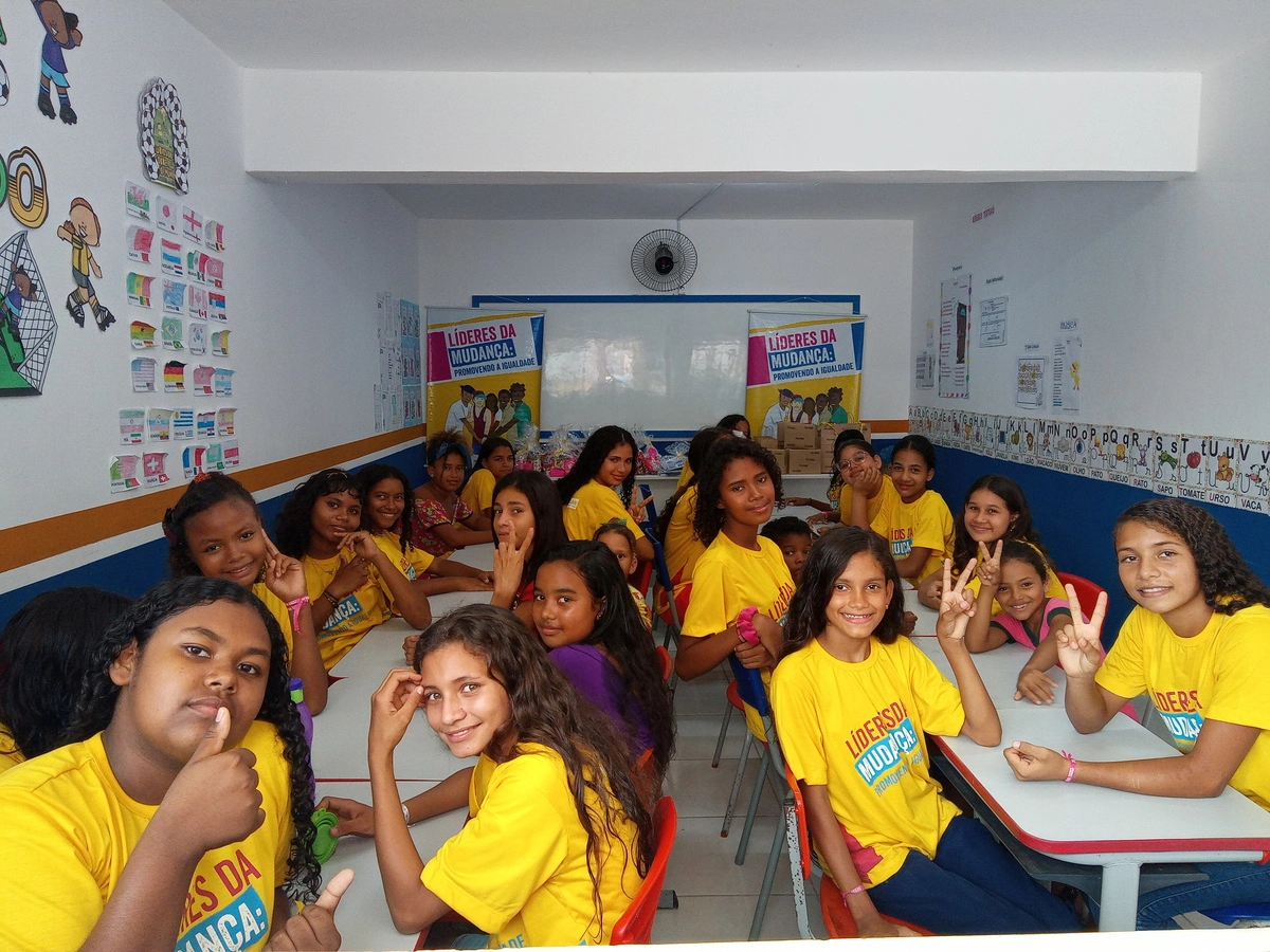 Turma de meninas em sala de aula, sorrindo e fazendo gestos de paz, durante atividade educativa do Projeto Líderes da Mudança.