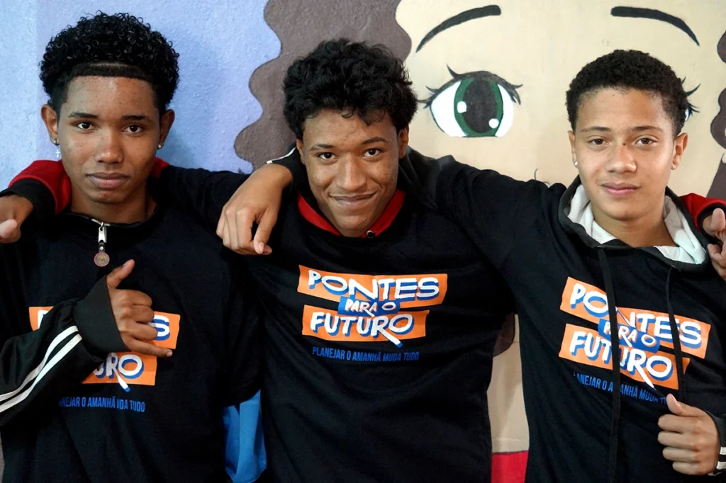 Três jovens com camisetas do projeto Pontes para o Futuro sorrindo
