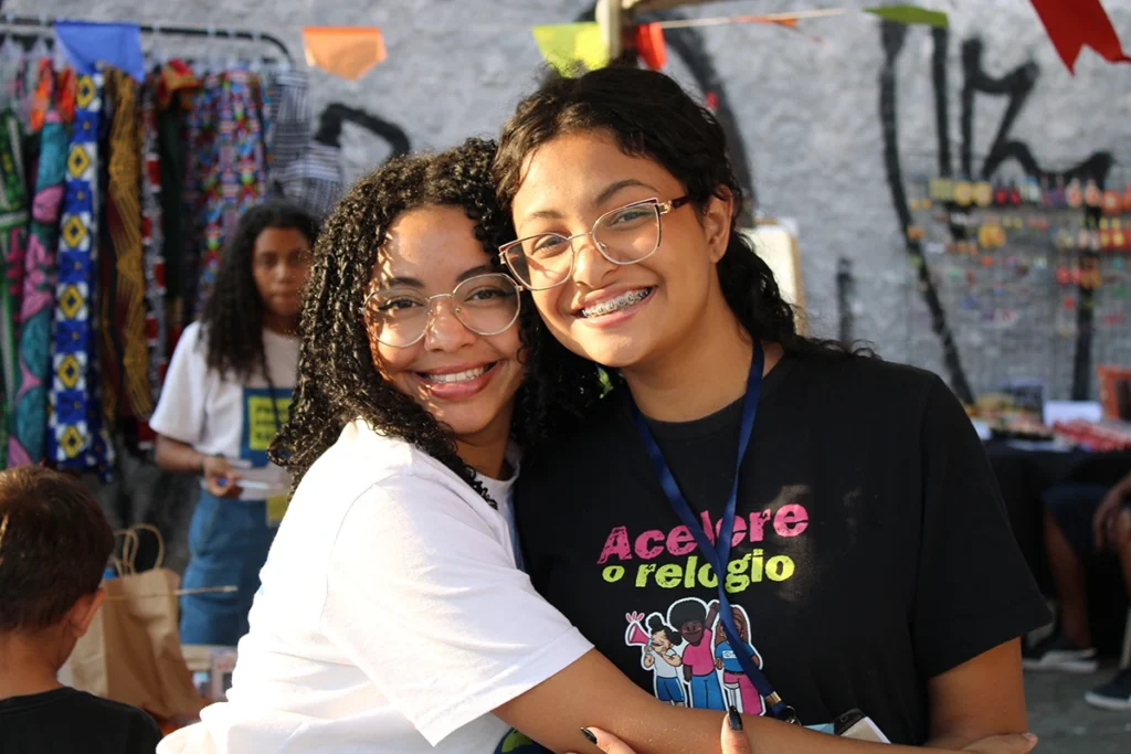 Duas adolescentes sorridentes abraçadas em evento comunitário, representando o espírito de amizade e empoderamento do Programa Adolescente Saudável.