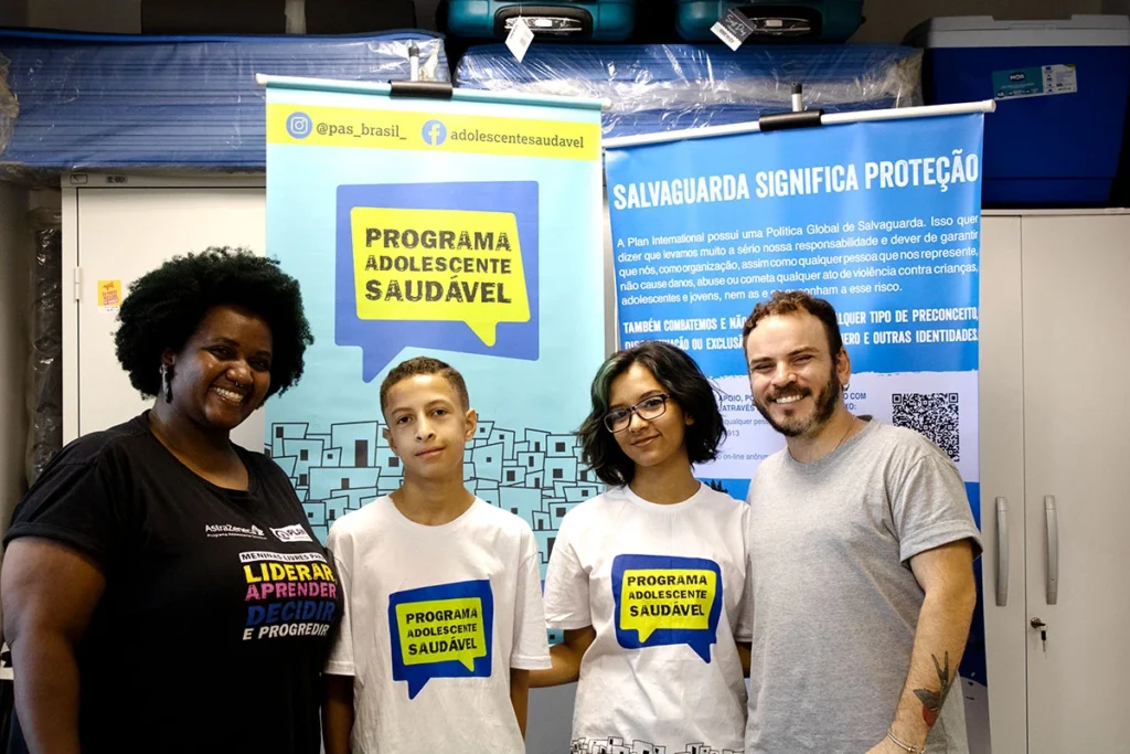 Educadora e dois adolescentes posam sorrindo em frente a banner do Programa Adolescente Saudável, que promove saúde e bem-estar na adolescência.