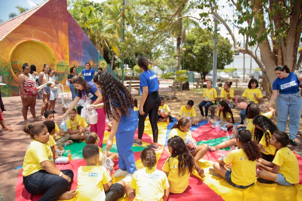 Crianças e educadoras sentadas em tapetes coloridos em uma praça, participando de dinâmica do Projeto Cambalhotas da Plan International Brasil.