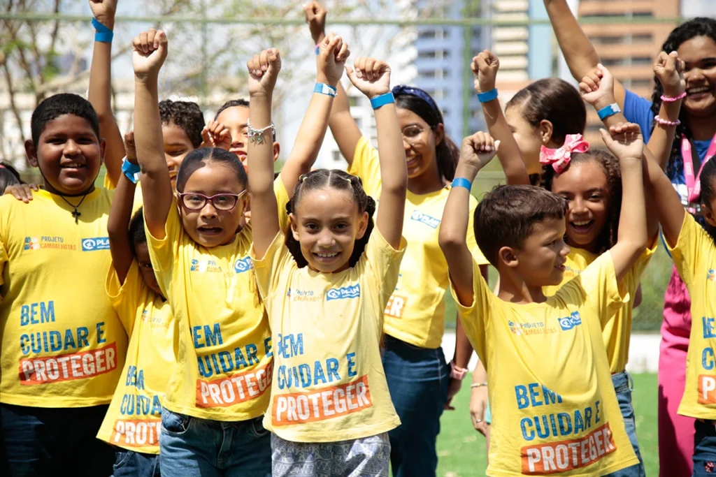 Grupo de crianças sorrindo com os braços erguidos, usando camisetas amarelas com a frase "Bem Cuidar e Proteger", em ação do Projeto Cambalhotas.