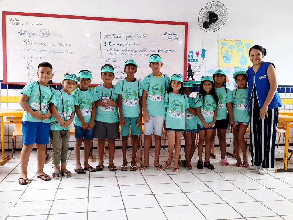Crianças uniformizadas com camisetas verdes e bonés participando de ação educativa do Projeto Comunidades Pelo Clima em sala de aula.