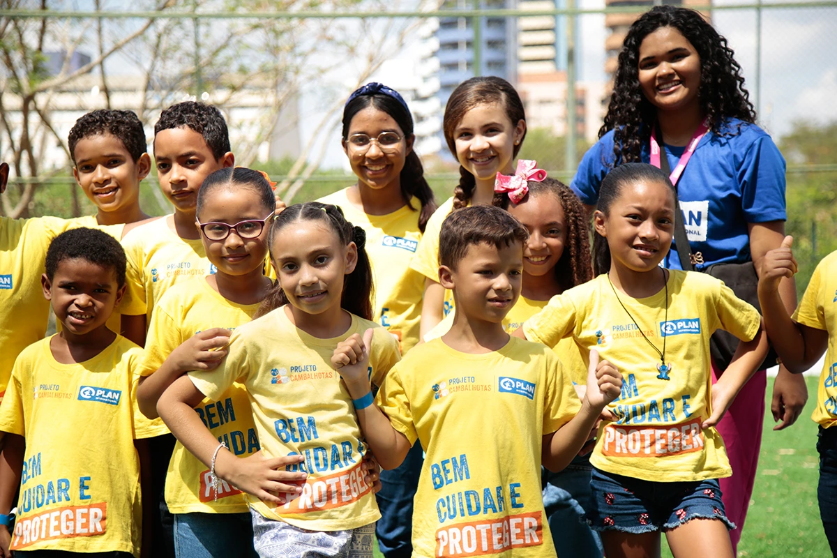 Crianças e educadoras posando juntas e sorrindo durante ação do Projeto Cambalhotas, com camisetas amarelas da campanha "Bem Cuidar e Proteger".